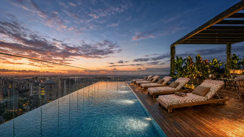 Piscina de borda infinita na cobertura ao anoitecer, com guarda-corpo de vidro, espreguiçadeiras em zigue-zague no deck de madeira, pergolado com plantas e a cidade iluminada ao fundo.