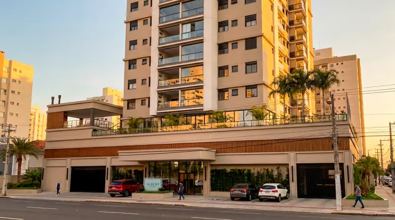 Torre residencial de fachada clara com sacadas de vidro sobre embasamento com faixa de madeira, palmeiras no terraço, carros em frente e pedestres na calçada ao fim da tarde.