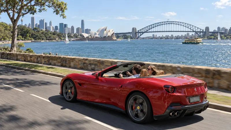 Casal num conversível vermelho passando pela estrada à beira da baía, com muro de pedra, a ponte em arco, a ópera e os prédios de Sydney ao fundo em dia de sol.