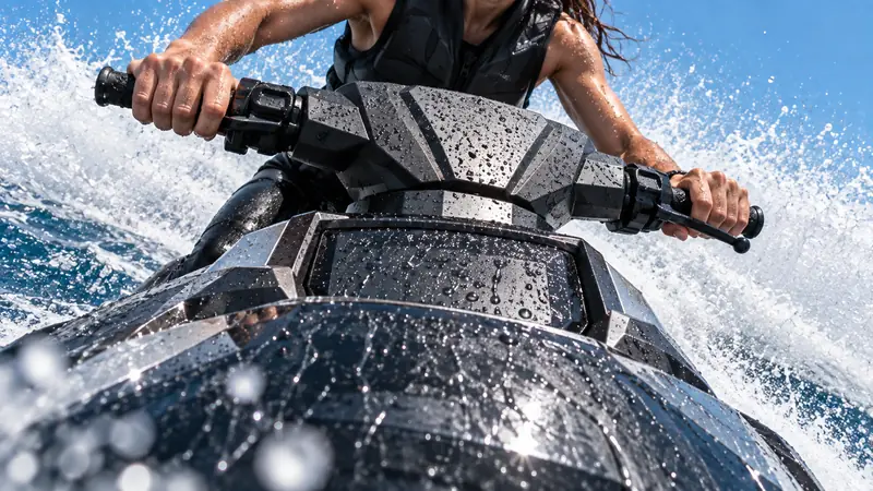 Frente de um jet ski preto e prata coberta de gotas, com as mãos do piloto no guidão e a água espirrando branca contra o céu azul.