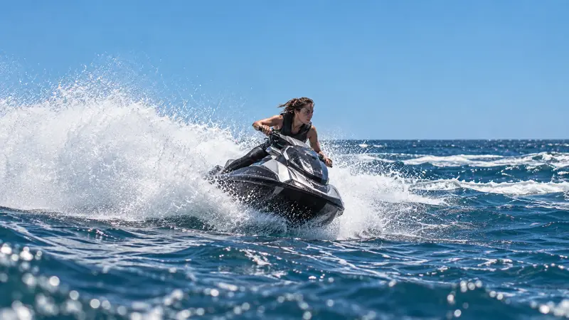 Mulher pilotando um jet ski escuro em curva, levantando uma cortina de água branca sobre o mar azul, com gotas brilhando em primeiro plano.