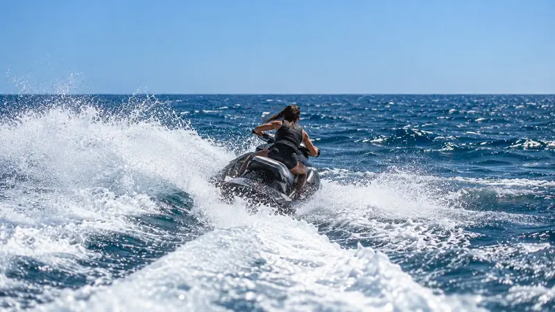Mulher de costas num jet ski afastando-se mar adentro, com o rastro de espuma em primeiro plano e o horizonte sob céu limpo.