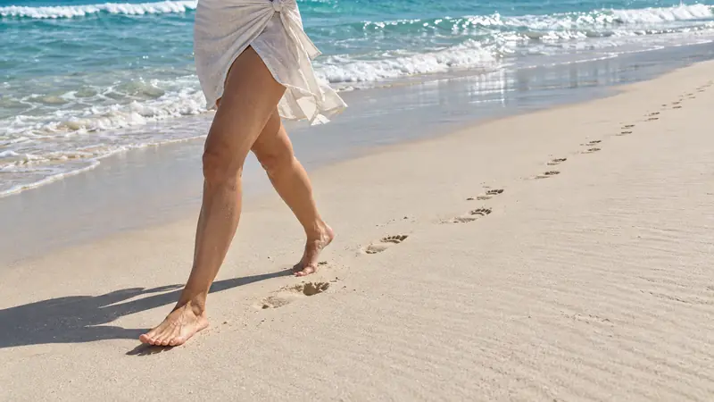 Close das pernas de uma mulher de saída de praia branca caminhando descalça na areia molhada, com a trilha de pegadas e a espuma das ondas ao lado.
