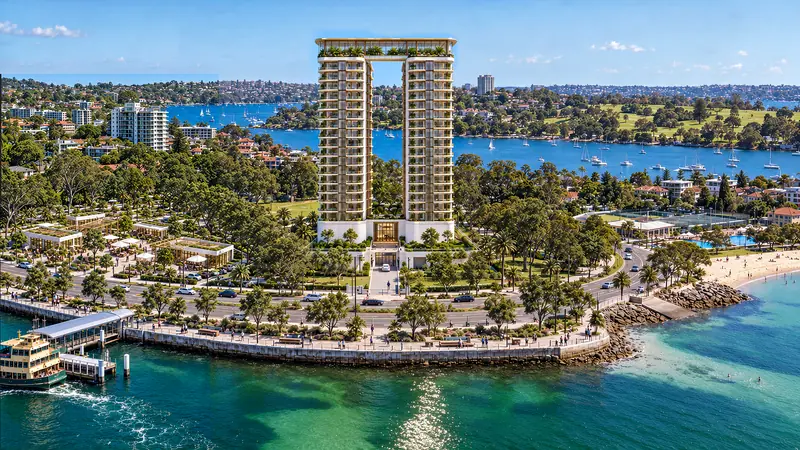 Duas torres claras unidas no topo por uma cobertura ajardinada, vistas do alto em dia de sol, numa península arborizada com cais de balsa, praia de água turquesa e a baía com veleiros ao fundo.