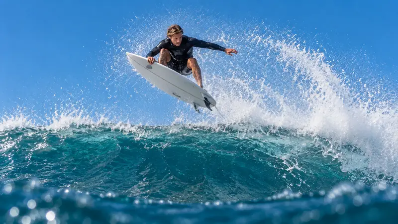 Surfista de roupa de borracha escura no ar sobre a crista da onda, com a prancha branca e um leque de água contra o céu azul.