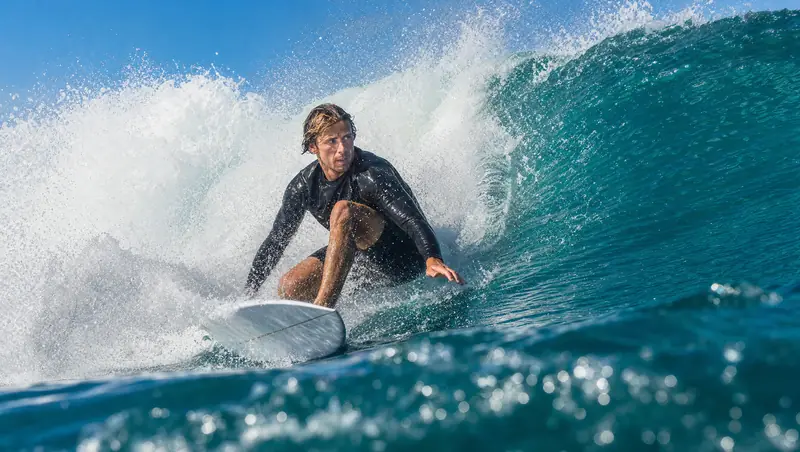Surfista agachado na prancha branca descendo a parede de uma onda verde-azulada, com a crista quebrando em espuma atrás dele.