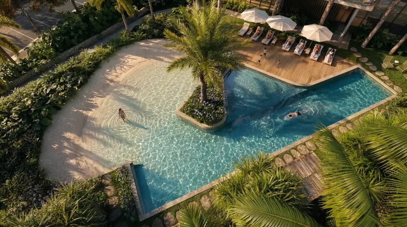 Piscina em L vista do alto, com prainha de areia, ilha com palmeira, raia para nadar, deck com espreguiçadeiras e guarda-sóis e jardins tropicais em volta.