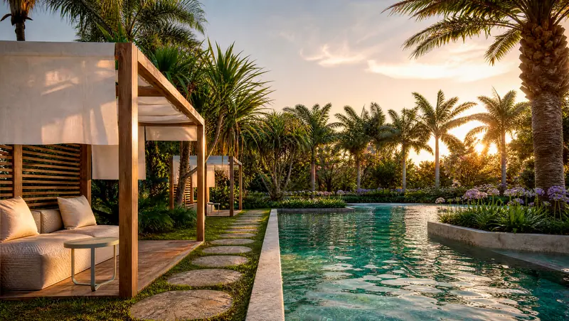 Piscina longa entre jardins tropicais ao pôr do sol, com cabanas de madeira e cortinas brancas, caminho de pedras no gramado e palmeiras ao redor.
