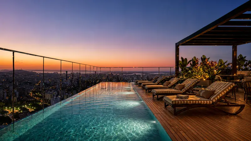 Piscina de borda infinita na cobertura ao anoitecer, com guarda-corpo de vidro, deck de madeira com espreguiçadeiras listradas, pergolado com plantas e cidade iluminada abaixo.