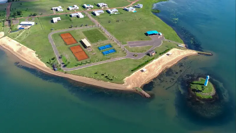 Península vista de drone com quadras de saibro, quadras azuis e campo gramado entre casas, faixa de areia contornando a água e um pequeno farol numa ilhota.