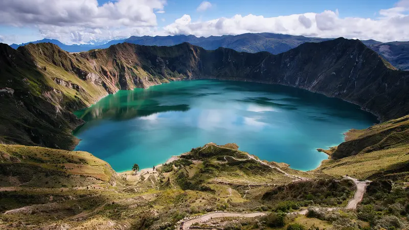 Lagoa de cratera de água turquesa vista do alto da borda em dia de nuvens altas, cercada por encostas íngremes de rocha e pasto e pela cordilheira azulada no horizonte.