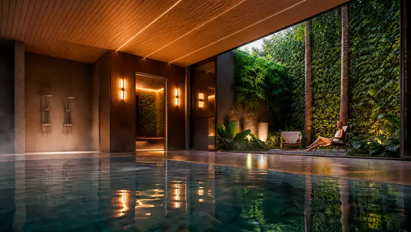 Piscina interna revestida de pedra escura sob forro de madeira com fitas de luz, arandelas e duchas na parede, aberta para jardim vertical onde uma mulher descansa numa cadeira.