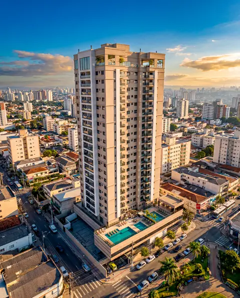 Torre residencial de fachada clara vista do alto no fim da tarde, com piscinas no terraço do embasamento, ruas arborizadas e cidade densa ao fundo.