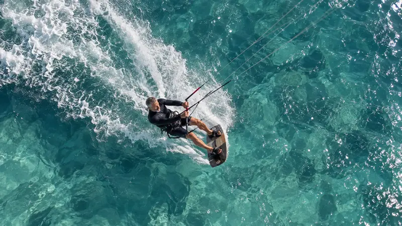 Kitesurfista fotografado de cima cortando água turquesa transparente sobre fundo de pedras, deixando rastro de espuma branca e com as linhas da pipa esticadas na diagonal.